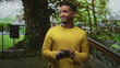 © Krakenimages.com - Young black man holding smartphone, thumbs typing while looking down, on a street beside a large tree and wooden stair railing; quiet contentment.