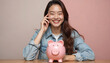 © Viktor - Happy young Asian woman smiles broadly, sitting at wooden table with pink piggy bank. Saves money for future, showing good financial planning, smart investment. Girl cheerful, thinking about wealth