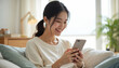 © Viktor - Young Asian woman smiles, relaxes on sofa using smartphone. She scrolls feed, chats online, enjoys leisure time at home. Modern life connect via mobile tech, peaceful atmosphere.