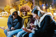 © Xavier Lorenzo - Multi-ethnic friends enjoying a winter night out, gathered on a bench, sharing laughs while browsing their smartphones under festive lights