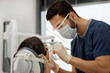 © Seventyfour - Young adult male dentist wearing protective mask and face shield treating patient in dental office, using dental instruments while focused on dental procedure, patient reclining in dental chair