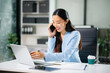 © laddawan - Asian Business woman Talking on the phone and using a laptop with a smile while sitting at office