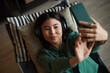 © DragonImages - Portrait of young adult Asian woman lying on couch wearing headphones holding smartphone taking selfie smiling slightly relaxed expression modern office environment