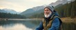 © Maryna - Elderly man with white beard and gray hat smiles near lake. He wears puffer vest, enjoys nature. Mountains and forest background. Active senior retiree.
