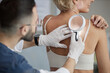 © Seventyfour - Caucasian middle aged woman undergoing skin examination by male dermatologist using magnifying glass, doctor wearing gloves closely inspecting patients back for dermatological assessment