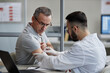 © Seventyfour - Middle aged Caucasian man receiving skin examination from young adult male dermatologist wearing gloves, sitting in medical office, patient looking at exposed arm