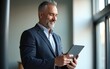 © ye - Happy middle aged business man ceo wearing suit standing in office using digital tablet. Smiling mature businessman professional executive manager looking away thinking working on tech device.