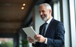 © ye - Happy middle aged business man ceo wearing suit standing in office using digital tablet. Smiling mature businessman professional executive manager looking away thinking working on tech device.