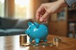 © Vadym - Hand puts gold coin into blue piggy bank on wooden table. Stacks of cash surround money box. Cozy room with soft light. Saving money for future goals.