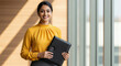 © PRASANNAPIX - Young Indian businesswoman in a yellow blouse confidently holding a black portfolio in a bright modern office corridor.