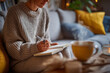 © Vitali - A woman journaling in a notebook on the couch with a steaming cup of tea.