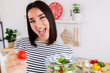 © Xavier Lorenzo - Young beautiful millennial girl eating healthy fresh green salad sitting in the kitchen, holding a fork with a cherry tomato