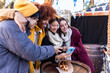 © Xavier Lorenzo - Group of multi-ethnic friends looking at photos on a smartphone at christmas market, hugging, smiling, and having fun together