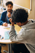 © Maskot - Rear view of tensed curly haired teenage boy leaning on elbow while sitting with counselor in high school office