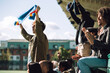 © Maskot - Young man cheering while holding sports scarf with raised hands at stadium stand on sunny day