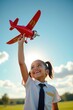 © Leonardo - Una niña vestida de piloto levanta con confianza su avión de juguete hacia el cielo
