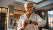 © photostockatinat - Senior Woman Checking Smartwatch: An elegant senior woman with short gray hair and glasses is focused on her smartwatch, checking information as she stands in a well-lit, modern living space.