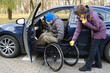 © Alex - A woman holds a wheelchair with her right hand, a male driver transfers into a wheelchair from his car