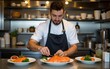 © li - Concentrated male cook in uniform standing at metal counter in kitchen of cafe and preparing delicious dish with salmon fish. High quality