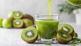 Green kiwi juice streams into a clear drinking glass, with whole and halved ripe kiwi fruits arranged around the base on a bright white surface, emphasizing freshness and healthy living