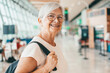 © luciano - Smiling senior woman in airport departure area looking at camera while waiting to board flight. Travel and tourism concept, elderly people with backpack