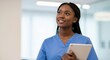 © Vasiliy - Young African American nurse in blue scrubs holding a tablet and looking up with a hopeful smile. Female medical professional in a modern hospital corridor.