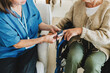 © shurkin_son - Image of two unrecognizable people, nurse in blue uniform doing saturation assessment and checking pulse with finger oximeter to check oxygen level of elderly woman in wheelchair at geriatric clinic