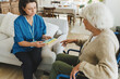 © shurkin_son - Upper view indoor image of nurse in blue uniform sitting on sofa in front of senior lady with physical disability in wheelchair and holding pill box, giving instructions of medication intake