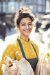 © Kowit - A smiling woman in a yellow outfit holds bags of groceries in a bustling outdoor market, radiating warmth and positivity.