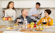 © JackF - Friendly elderly parents enjoying family dinner with young adult son and daughter-in-law, sharing stories and laughing over wine and light snacks in cozy kitchen