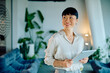 © we.bond.creations - Confident Asian businesswoman holding a digital tablet with a happy expression, standing in a contemporary office setting
