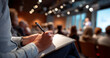© perfectlab - Close-up of person taking notes with pen on notebook during a conference or seminar in a modern meeting room with blurred audience and speaker