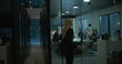 © Marco - Female executive entering glass boardroom as colleagues sit around table with laptops during late office meeting in modern corporate workspace