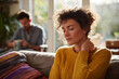 © EricMiguel - Young woman in a cozy yellow sweater rubbing her neck on a sunlit sofa at home, tired and pensive as partner sips coffee