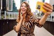 © maxbelchenko - Selfie time! Young woman is enjoying herself with phone in bustling cityscape. Beautiful tourist enjoys stroll, blogs on sunny day against backdrop of metropolis. Blogging, urban, tourism concepts.