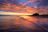 Beautiful sunrise reflections on coast at high tide with Bamburgh Castle in distance. Northumberland, North East England, UK.