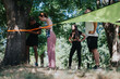 © qunica.com - A diverse group of friends or teammates participates in an outdoor fitness activity, tying an orange strap to a tree and standing under a green shade sail in a sunny park.