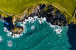 © AmazingAerialAgency - Aerial view of rugged cliffs meet the turquoise sea, where white waves crash against the dark rocks beneath a vibrant green coastline, St. Davids, Wales, United Kingdom.