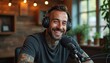 © Pete - Smiling man with tattoos wearing headphones. He sits at microphone recording podcast in cozy studio. Broadcaster talks into mic. Audio equipment setup for live show or interview.