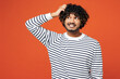 © ViDi Studio - Young sad unhappy disappointed mistaken Indian man he wears striped sweatshirt casual clothes hold scratch head look aside isolated on plain red orange background studio portrait. Lifestyle concept.