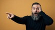 © David - Shocked bearded man with wide eyes points his finger to the side with an open mouth in disbelief, holding his head in amazement on a solid yellow background