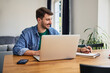 © baranq - Young man at home sitting at dinning table doing notes while working with laptop