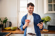 © baranq - Young smiling man using mobile phone while drinking morning coffee at home