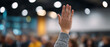 © perfectlab - Raised hand of a person in a gray sweater during a meeting or conference with blurred audience and bokeh lights in the background