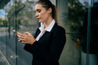 © BullRun - Young woman in business suit standing near office glass, focused on smartphone, embodying digital independence, mobile productivity and freelance mindset.