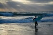 © ELENI MAVRANDONI - Seascape with scenic view of a male surfer on a wet suit walking with his surfboard into the surf, at time of sunset in Oceanside Beach, San Diego Southern California.