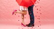 © Vasiliy - A romantic couple kissing under a red umbrella with falling heart confetti. Cropped view of a man and woman's legs on a pink background for Valentine's Day