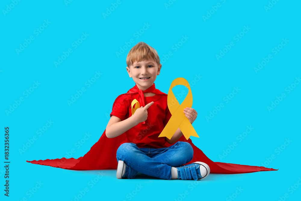 Little boy dressed as superhero pointing at gold ribbon on blue background. Childhood cancer awareness concept