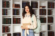 © Pixel-Shot - Female student with backpack holding stack of books near bookshelves in library
