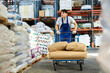 © JackF - Young male employee is driving trolley cart with several bulky paper bags. Guy works as loader in a warehouse of construction materials